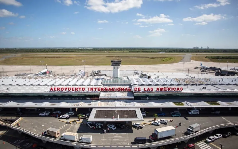 Vista frontal del aeropuerto Las Américas (SDQ), con el nombre de la terminal, torre de control y aviones al fondo.